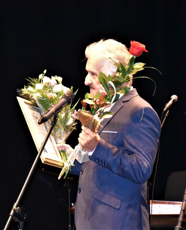 Przy mikrofonie stoi Laureat Angelusa animatora kultury – Aldon Dzięcioł. W prawej ręce trzyma Dyplom-Certyfikat i wiązankę kwiatów, w lewej statuetkę i czerwoną różę. Zadumany, ale uśmiechnięty dziękuje Kapitule za nagrodę.