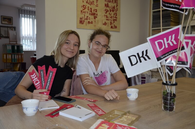 W pomieszczeniu czytelnianym przy stoliku siedzą dwie uśmiechnięte dziewczyny. Pozują do zdjęcia. Pierwsza z lewej, trzyma w dłoni różowe zakładki do książek z logo DKK. Na stoliku przed nimi stoją filiżanki z napojem, leżą notesiki z logo DKK. Po prawej stronie, na stole stoi flakonik z różowymi i białymi chorągiewkami DKK