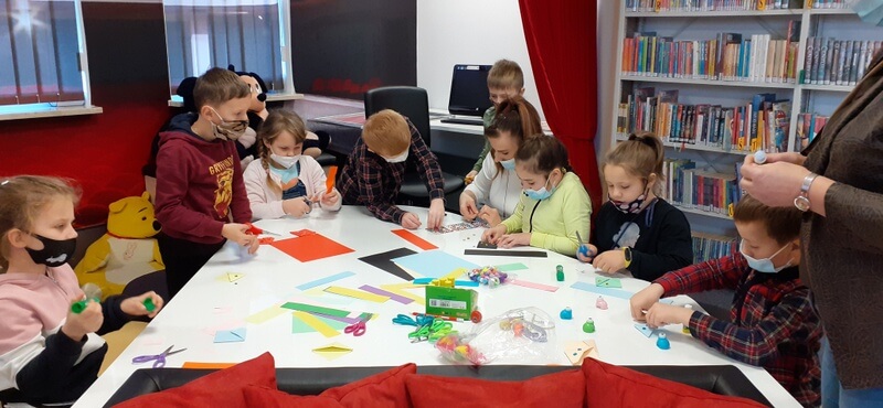 Oddział dla Dzieci, uczniowie i panie nauczycielki siedzą dookoła białego stołu, na którym leżą kolorowe kartoniki, kleje, nożyczki, piórka, pomponiki i inne barwne ozdoby. Dzieci przygotowują zakładki metodą origami. W tle po prawej stronie widnieje wysoki regał z kolorowymi książkami.