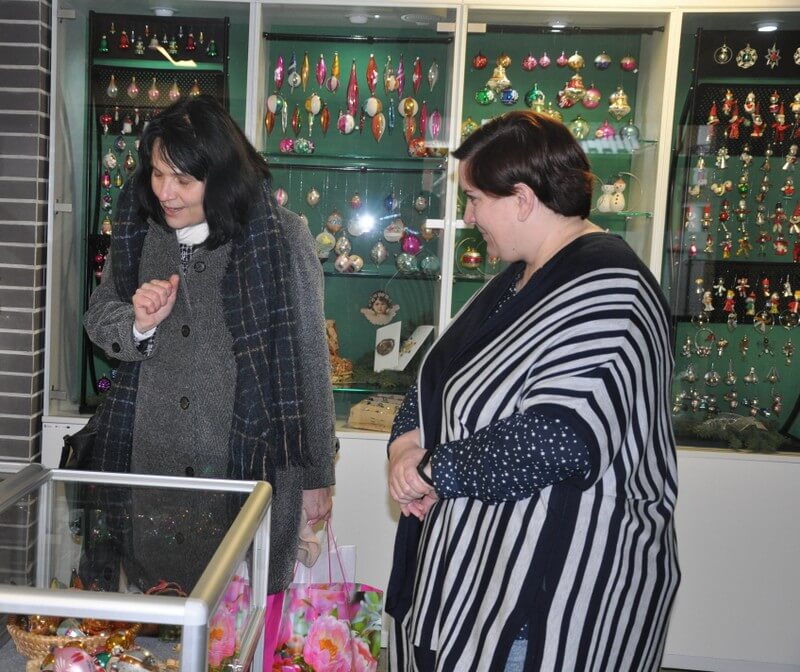 Na pierwszym planie dwie kobiety, poseł Anna Dąbrowska-Banaszek i autorka wystawy, oglądają dekoracje świąteczne w stojącej oszklonej gablocie, w tle oszklona 4-dzielna gablota z wiszącymi ozdobami świątecznymi