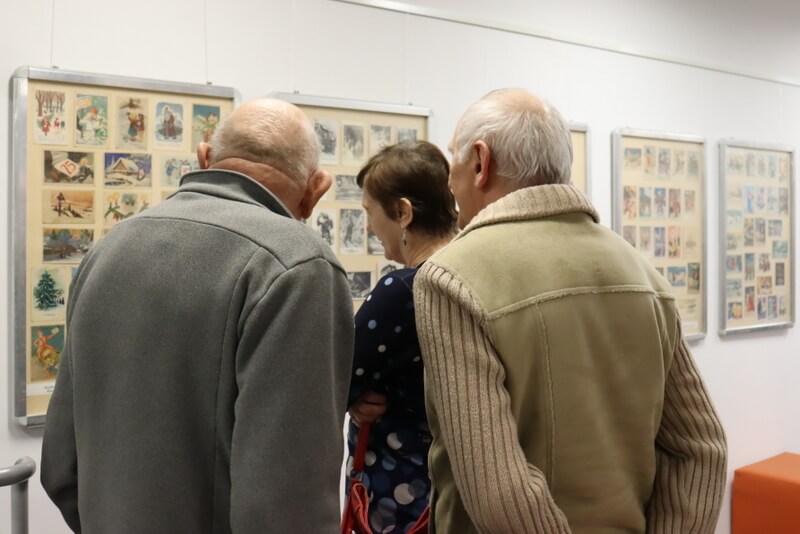 Trójka ludzi ogląda świąteczne kartki pocztowe eksponowane w antyramach zawieszonych na ścianach. 