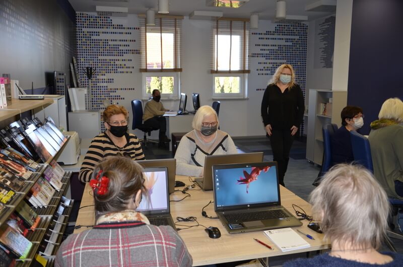 Uczestnicy siedzą przy komuterach, prowadząca stoi z prawej strony. 