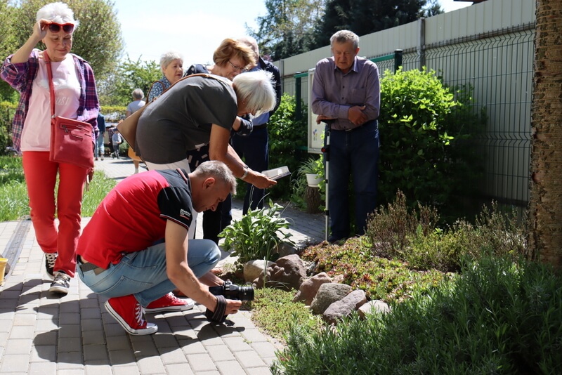 Prowadzący i uczestnicy robią zdjęcia roślinom w bibliotecznym ogródku. Kilku uczestników przygląda się robiącym zdjecia. 