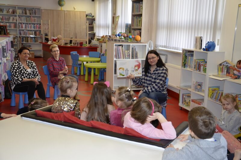 Oddział dla Dzieci. Przedszkolaki siedzą na kanapie przy białym stole, w tle wnętrze Oddziału dla Dzieci, czerwone biurko, regały z książkami. W centrum zdjęcia prowadząca (J. Orzeszko) pokazuje ilustracje z książki. Po lewej stronie nieco w głębi sali na niebieskich krzesełkach siedzą dwie panie nauczycielki, uśmiechają się do prowadzącej i do dzieci.
