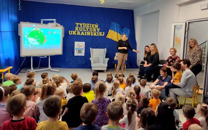Na pierwszym planie widzimy grupę dzieci skierowanych twarzami ku pani dyrektor przedszkola, która stoi w oddali i przedstawia siedzących gości po jej lewej stronie. Tło jest w kolorze niebieskim, na którym widnieje napis TYDZIEŃ KULTURY UKRAIŃSKIEJ. Po prawej stronie zawieszona jest flaga ukraińska, zaś po lewej plakat Mała Książka – Wielki Człowiek, przy którym stoi projektor.