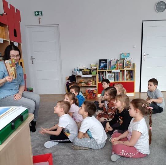 Widok z boku, na pierwszym planie, na szarej wykładzinie siedzi po turecku grupka uśmiechniętych dzieci i przygląda się prezentowanej przez czytającą książeczki. W tle regalik z książkami o żywiołach Ziemi. Z lewej strony siedzi czytająca - Joanna Orzeszko i pokazuje dzieciom okładkę książki.