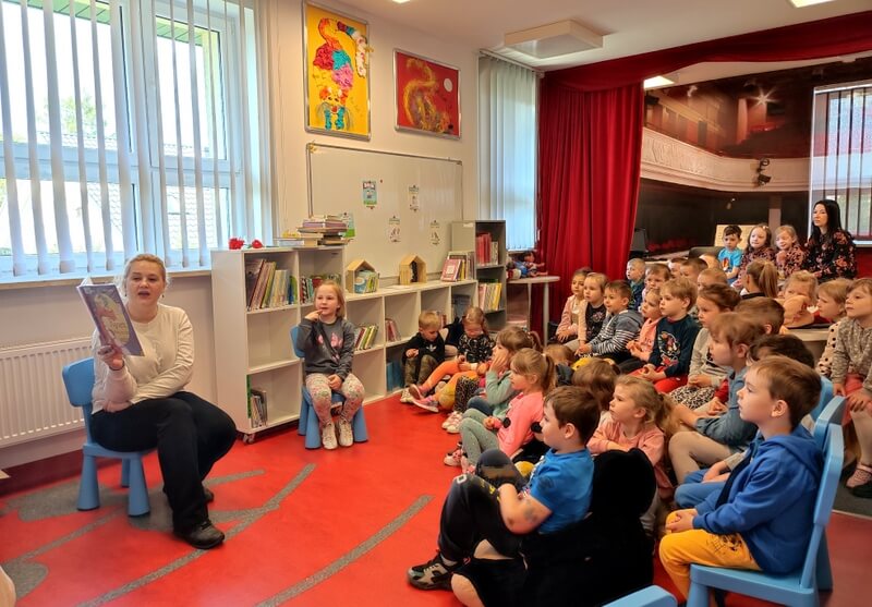 Z prawej strony siedzi grupa dzieci skierowana twarzami do prowadzącej zajęcia, która czyta dzieciom książkę. Tło stanowi wystrój biblioteki w barwach czerwono czarnych oraz regały z książkami.