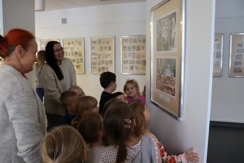 Galeria Na Parterze, grupa dzieci ogląda plansze ze zdjęciami ludzi. Plansze eksponowane są na ruchomych ściankach systemu wystawienniczego. W tle, na białych ścianach wiszą plansze z kolekcją kart świątecznych.