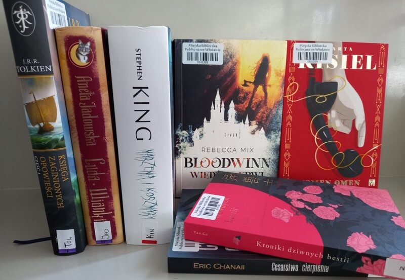 Trzy książki stojące, widoczne grzbiety, dwie książki stojące widoczne okładki: Rebecca Mix: Bloodwinn. Wiedźmy krwi i Marta Kisiel: Nomen omen. Dwie książki leżące, widoczne grzbiety.