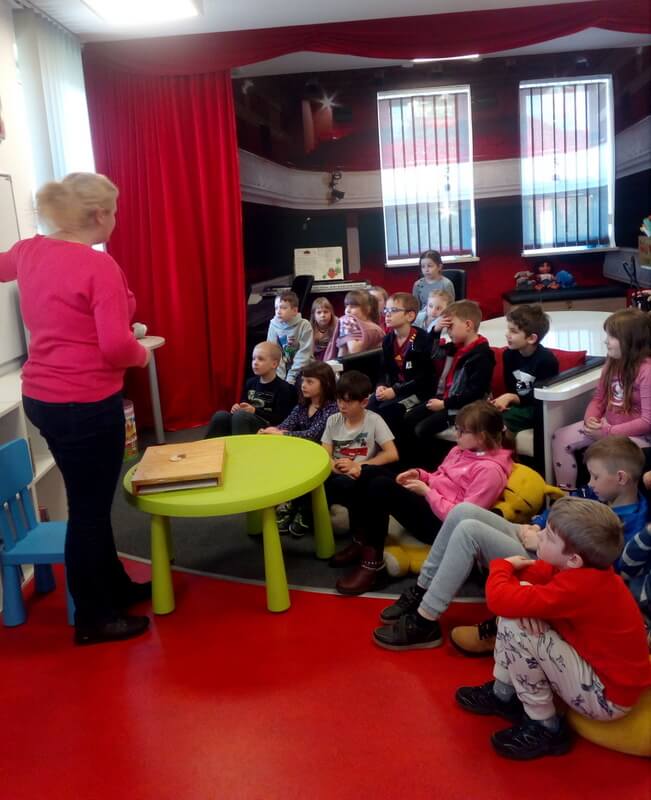 Prowadząca zajęcia stoi przed grupą dzieci, które skierowane są twarzami do niej. Tło stanowi wystrój biblioteki w kolorach czerwono – czarnych.