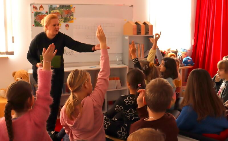 Na pierwszym planie widzimy grupę dzieci zgłaszających się do odpowiedzi. Po lewej stronie, stoi prowadząca zajęcia, która gestem dłoni wskazuje dziecko do odpowiedzi. Tło stanowi biała tablica z kolorowymi planszami pomocniczymi oraz regały z książkami.