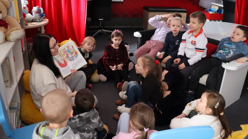 Grupka dzieci siedzi na białej kanapie w kształcie fortepianu, część dzieci  siedzi na pluszowych siedziskach (żółte misie i czarno- różowe myszki) w półkolu przed prowadzącą zajęcia. Prowadząca siedzi na szarej wykładzinie i pokazuje dzieciom obrazki z książki. Za plecami prowadzącej fragment niskiego regału z kolorowymi książkami, wyżej siedzi brązowy miś. W tle czerwona kurtyna.