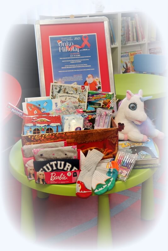 Na stoliczku wyeksponowany jest plakat promujący akcję Onko Mikołaj oraz pudełko z prezentami dla dzieci. Tło stanowi wystrój biblioteki w postaci regałów z książkami.
