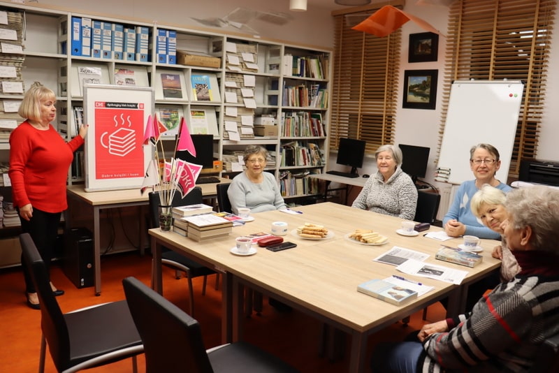 Grupa kobiet siedzi przy stole. Jedna z nich stoi i pokazuje plakat. Na stole stoją filiżanki, talerzyki z ciastkami, chorągiewki z logo DKK, obok  leży kilka książek. 