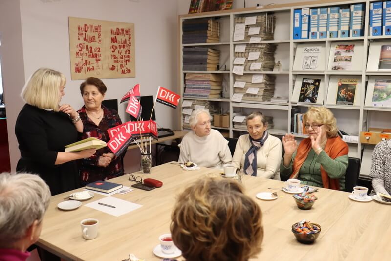 Spotkanie grupy kobiet. Kilka z nich siedzi przy stole, na którym stoją filiżanki i talerzyki z ciastem oraz chorągiewki z napisem DKK.Dwie z Pań  stoją, jedna trzyma w ręku książkę i tłumaczy coś drugiej