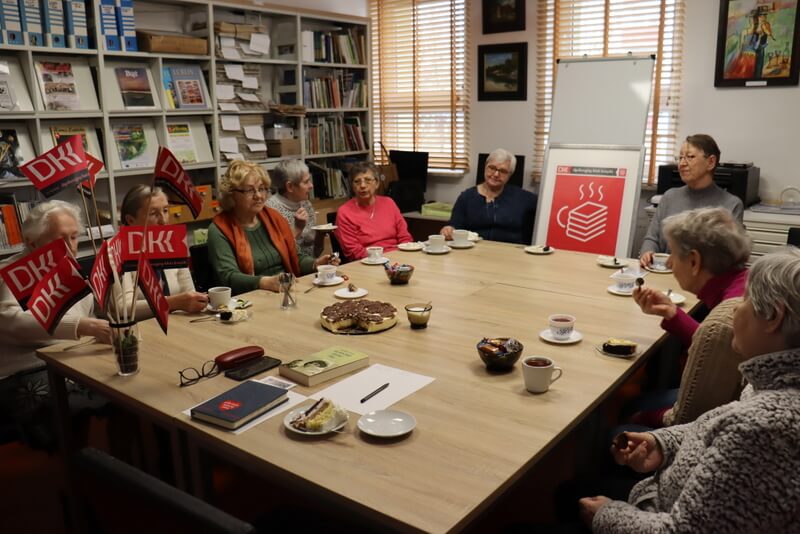 Grupa kilkunastu kobiet siedzi przy stole. Piją kawę i jedzą ciasto. Na stole leży książka, kartki, długopisy, stoją chorągiewki z logo DKK i duży plakat.W tle na regałach znajdują się książki i czasopisma