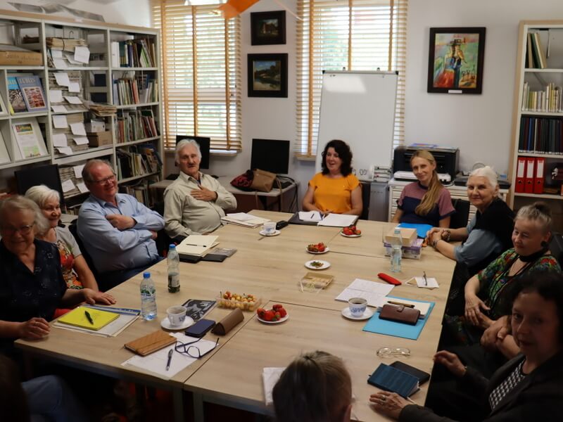 Grupa poetów siedzi przy stole. W tle z lewej strony regały z książkami i prasą, z prawej okno.