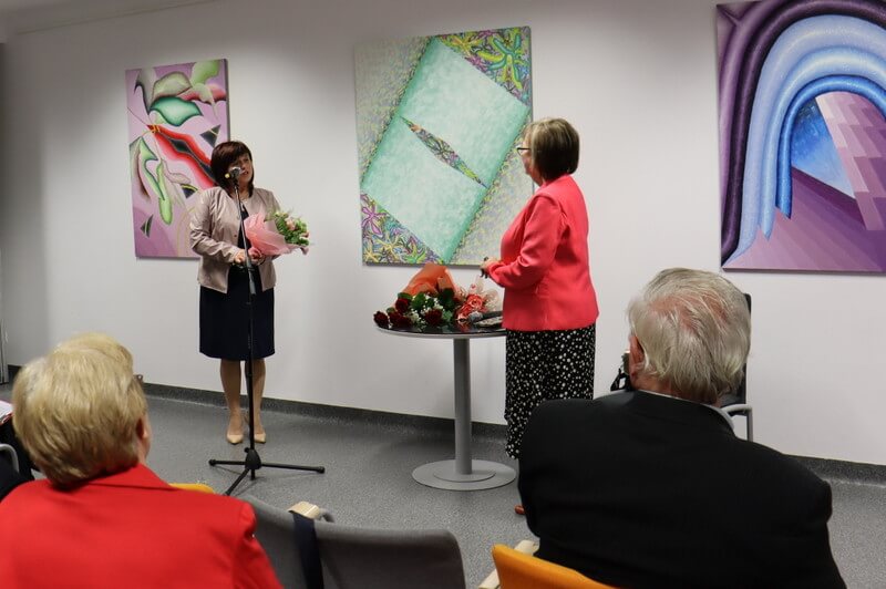 Dwie kobiety stoją przed ścianą z obrazami. Kobieta po lewej trzyma bukiet kwiatów