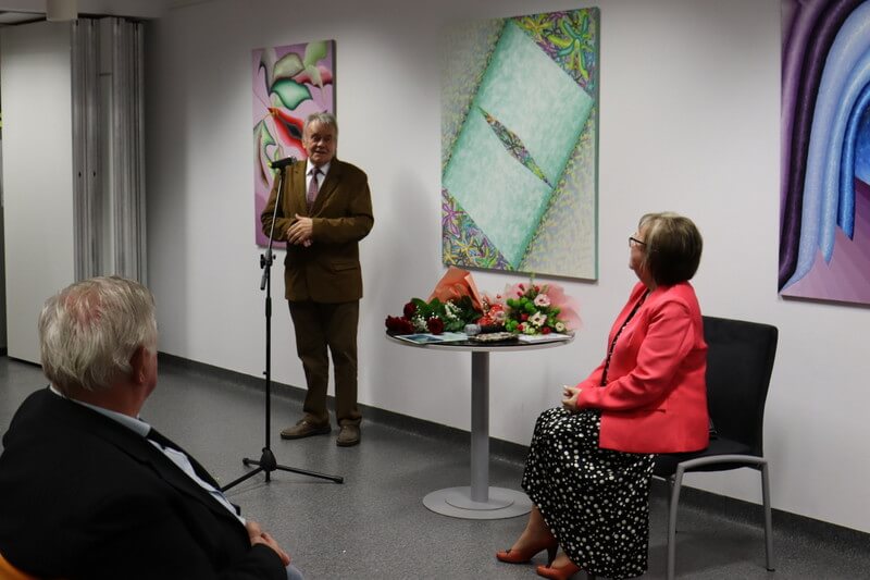 Dwoje ludzi przed ścianą z obrazami. Mężczyzna stoi przed mikrofonem, kobieta siedzi przy stoliku. 