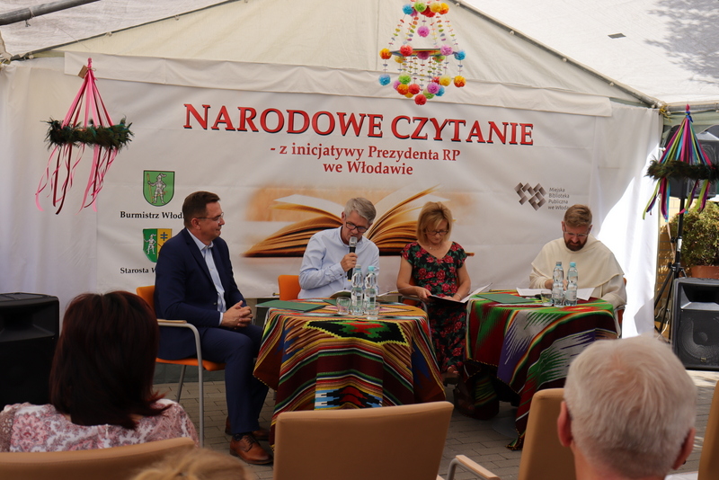 Grupa ludzi siedząca przy dwóch stolikach. Za nimi wisi baner - Narodowe Czytanie z inicjatywy Prezydenta RP we Włodawie. Jedna z osób czyta tekst do mikrofonu.