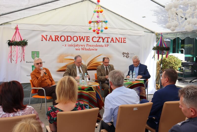 Grupa ludzi siedząca przy dwóch stolikach. Za nimi wisi baner - Narodowe Czytanie z inicjatywy Prezydenta RP we Włodawie. Jedna z osób czyta tekst do mikrofonu.