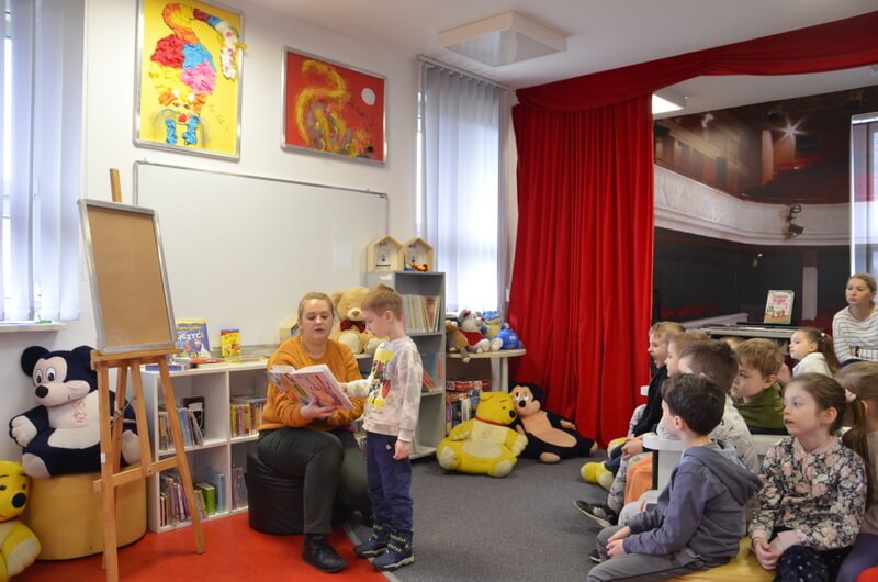 Z prawej strony grupa dzieci siedzi na pufach, skierowana twarzami do prowadzącej, która prezentuje książkę. Jeden z przedszkolaków stoi przed prowadzącą, pokazując dłonią obrazki w książce. Po lewej stronie stoi sztaluga na której wyeksponowane jest puste płótno. Tło stanowi wystrój biblioteki.