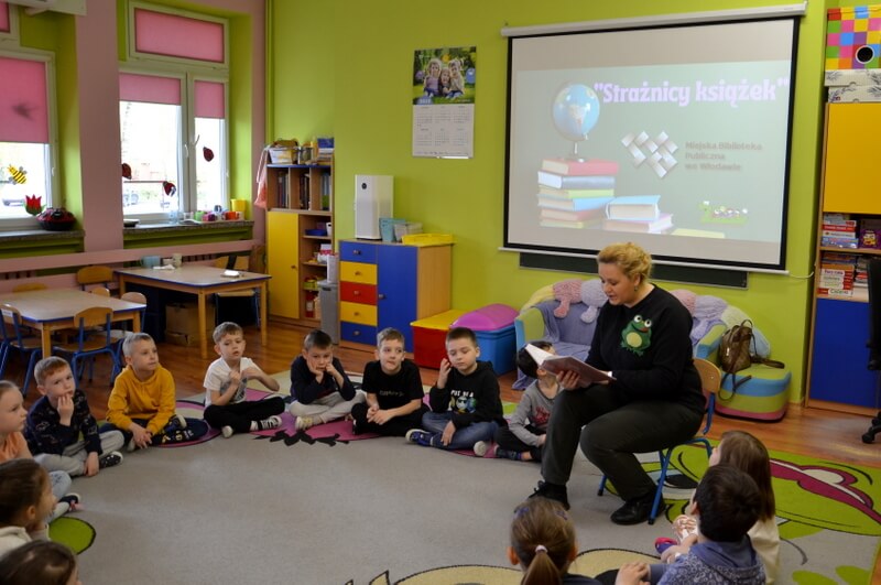Zaproszona bibliotekarka czyta dzieciom bajkę na dywanie. Dzieci wraz z nią tworzą okrąg. Tło stanowią kolorowe meble i zabawki przedszkolne. Za prowadzącą widnieje dekoracja w postaci książek i napisu: Strażnicy książek.