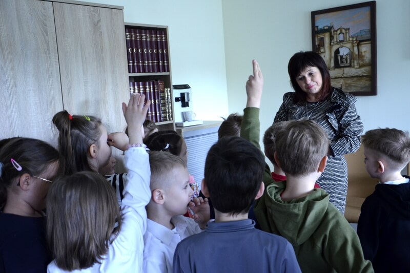 Grupa dzieci wraz z wychowawczynią są w gabinecie Pani Dyrektor – Miejskiej Biblioteki Publicznej we Włodawie. Dzieci zgłaszają się do odpowiedzi tło stanowią meble z książkami.