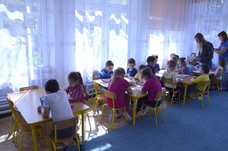 Dzieci w grupach siedzą przy stolikach i rysują. Opiekunowie grupy rozdają dzieciom kartki. 