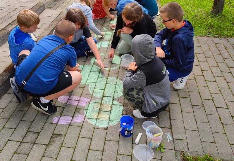Grupa dzieci maluje kredą na kostce brukowej dużą zieloną gąsienicę. Po prawej stronie leży na  kostce brukowej pojemnik z kolorową kredą.