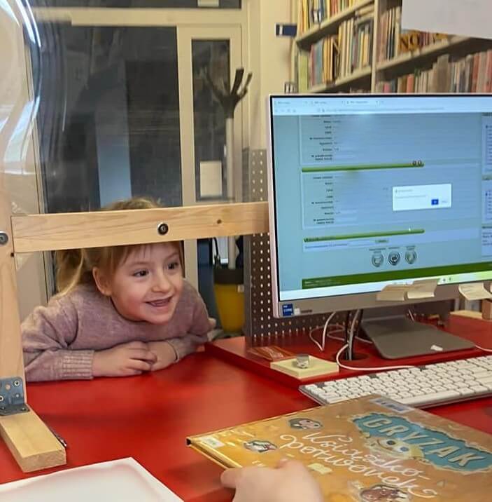 Oddział dla Dzieci. Widok zza biurka bibliotekarza. Na pierwszym planie fragment czerwonej lady bibliotecznej, ekran komputera i uśmiechnięte dziecko wypożyczające książkę. Dziecko uśmiecha się zaglądając pod pleksą zabezpieczającą stanowisko bibliotekarza – pozostałość po pandemii covid 19. 