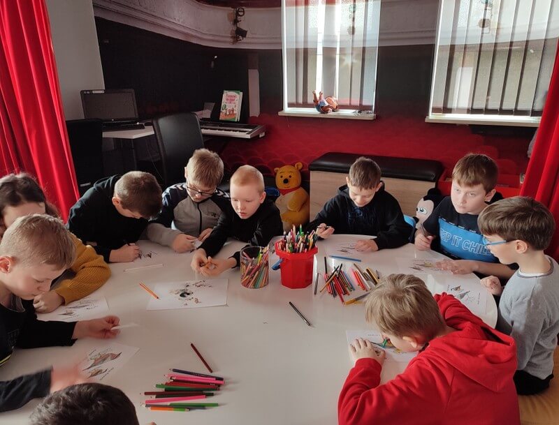 Grupa dzieci siedzi wokół stołu na którym tworzą prace plastyczne. Tło stanowi wystrój biblioteki w barwach czerwono - czarnych.
