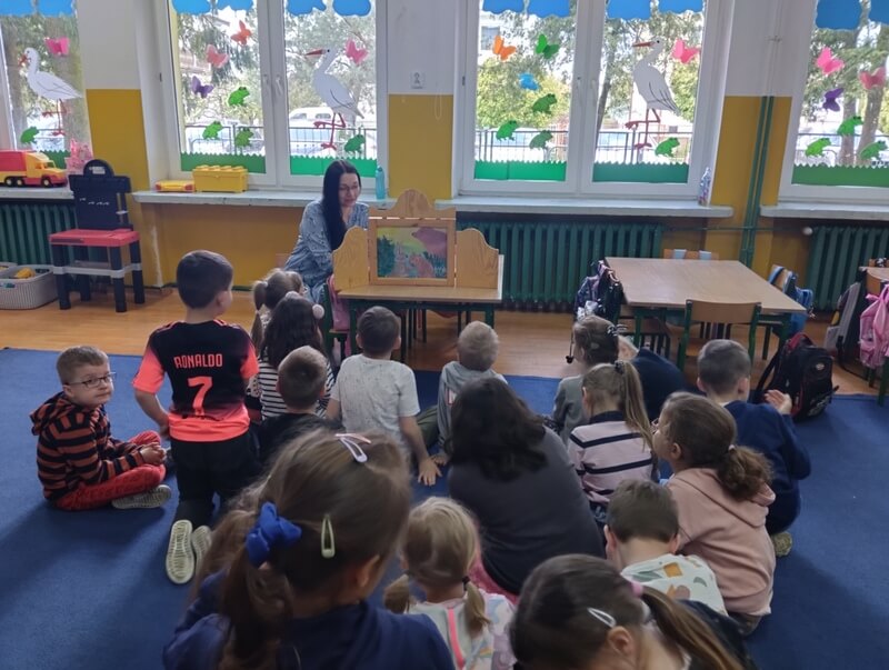 Na niebieskim dywanie siedzi grupa dzieci zasłuchanych w bajkę prezentowaną w teatrzyku Kamishibai. Prowadząca czyta bajkę, dzieci oglądają przesuwane, kolorowe plansze w drewnianej skrzyneczce (teatrzyku). W tle, za prowadzącą (J. Orzeszko) są dwa duże okna ozdobione wiosenną dekoracją. Motywy wiosenne: błękitne chmurki, bociany, żabki, kwiatki i motylki wśród zielonej trawy. Dekoracje wycięte są z kolorowych kartonów.