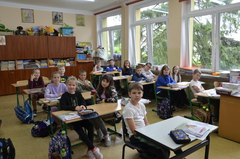 Uczniowie siedzą przy ławkach w klasie, skierowane twarzami do fotografa. Tło stanowią meble, książki i pomoce naukowe. 