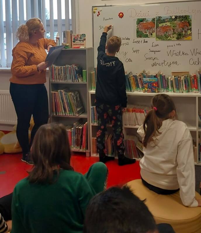W oddali stoi przy tablicy bibliotekarka wraz z uczniem. Uczeń pisze na tablicy odpowiedzi.  Wokół niego siedzi grupa dzieci. Tło stanowi wystrój biblioteki w postaci regałów z książkami.