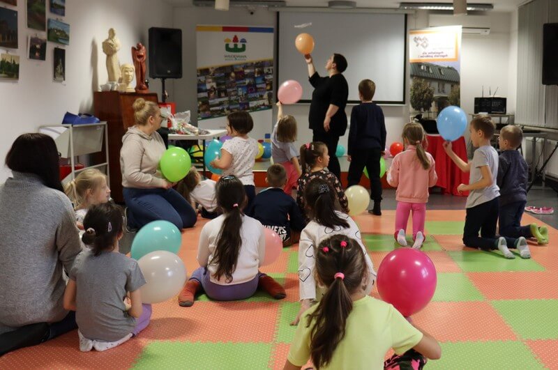 Grupa dzieci skierowana tyłem do osoby fotografującej bawi się kolorowymi balonami. W oddali pani Ewa prezentuje sztuczki dzieciom. Tło stanowią wyeksponowane prace plastyczne i plansze z logiem biblioteki.