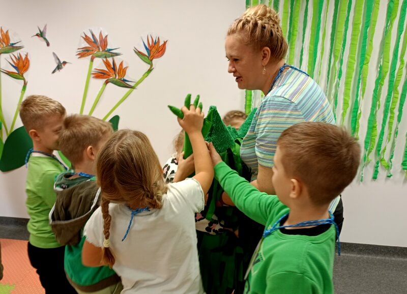 Bibliotekarka trzyma pluszowego Zielonego Potwora. Wokół nich stoi grupa dzieci, które „przybijają piątkę” pluszakowi.