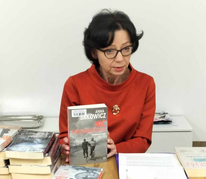 Pracownik biblioteki siedzi przy stole, w ręku trzyma książkę, widoczna okładka: Anna Sakowicz: Nić przeznaczenia.