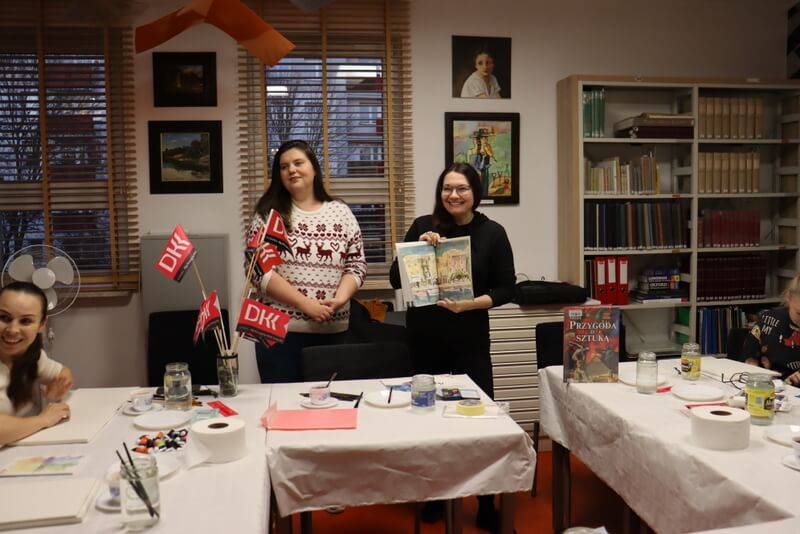 Czytelnia Działu Regionalnego. Prowadząca warsztaty (jasny sweter) i moderator Dyskusyjnego Klubu Książki (czarna bluza) otwierają zajęcia. Moderator trzyma w dłoniach książkę pokazuje młodym kobietom siedzącym przy stole. Stół nakryty białym, papierowym obrusem. Na stole przed każdą uczestniczką stoi słoik z wodą, pędzle, kolorowe farby i papier do malowania. W tle widoczne różowe chorągiewki z logo Dyskusyjnego Klubu Książki DKK, regały z książkami.