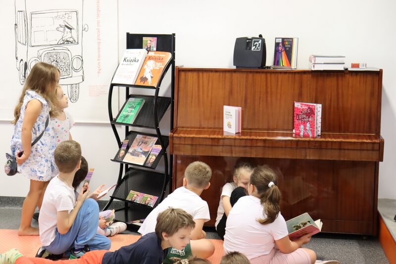 Grupa dzieci ogląda książki podczas spotkania w bibliotece. Część z nich siedzi na podłodze przy pianinie, na którym ustawiono kilka publikacji, a inne stoją przy stojaku z książkami