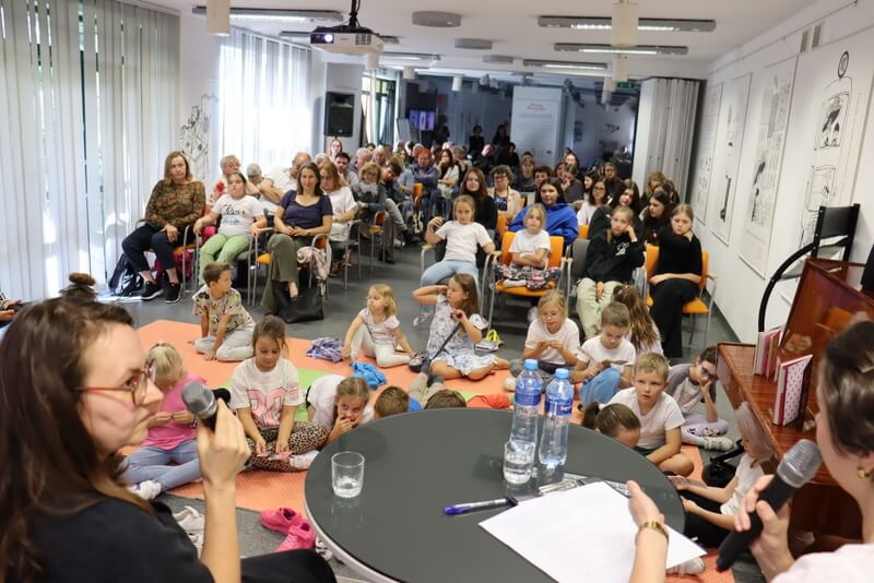 Spotkanie autorskie w bibliotece. Na pierwszym planie dwie osoby z mikrofonami prowadzą rozmowę przy stole z wodą i notatkami. Przed nimi na podłodze siedzi grupa dzieci, a za nimi na krzesłach liczna publiczność dorosłych i młodzieży. Sala jest jasna, z dużymi oknami po lewej stronie i ilustracjami na ścianach
