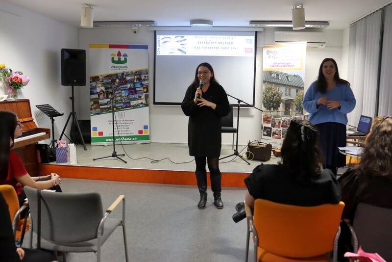 Sala Konferencyjna MBP we Włodawie. Zbliżenie na prowadzącą wernisaż J. Orzeszko, która przemawia do publiczności (poza kadrem). Po prawej stronie stoi uśmiechnięta artystka, K. Melcher. W tle wyświetlona na ekranie belka z informacją o wydarzeniu. Po lewej stronie na pianinie stoją wazony z kwiatami dla artystki.