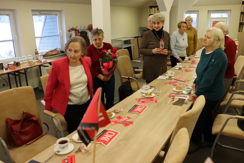 Grupa kobiet stoi przy stole. Jedna z nich trzyma w ręku bukiet kwiatów. Wszystkie zwrócone są w jednym kierunku, słuchają i uśmiechają się. Na stole leżą materiały promocyjne z logo DKK, stoją filiżanki i talerz z ciastem.