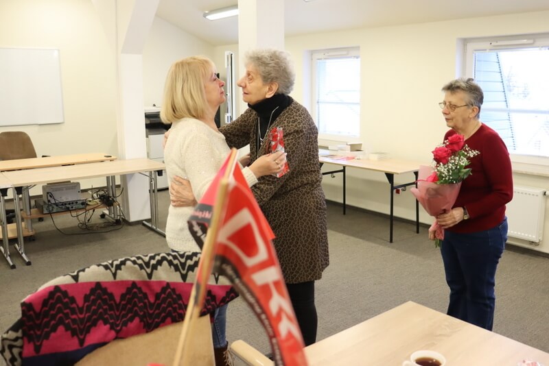 Trzy kobiety stoją na środku sali. Jedna z nich trzyma w ręku bukiet kwiatów. Dwie pozostałe zwrócone twarzą do siebie rozmawiają a jedna z nich trzyma coś w ręku.