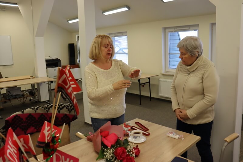 Dwie kobiety stoją przy biurku, na którym leżą kwiaty i stoi flakon z chorągiewkami z logo DKK. Jedna z nich pokazuje coś drugiej. W tle ściana i dwa okna.