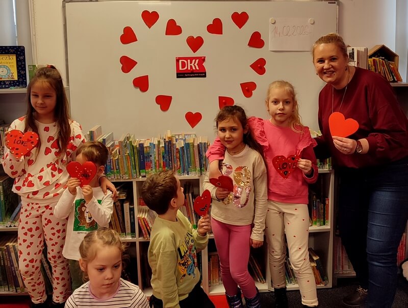 Grupa dzieci wraz z bibliotekarką trzymają w dłoni serduszka pozują do wspólnej fotografii. Tło stanowi dekoracja w postaci serduszka i loga DKK.