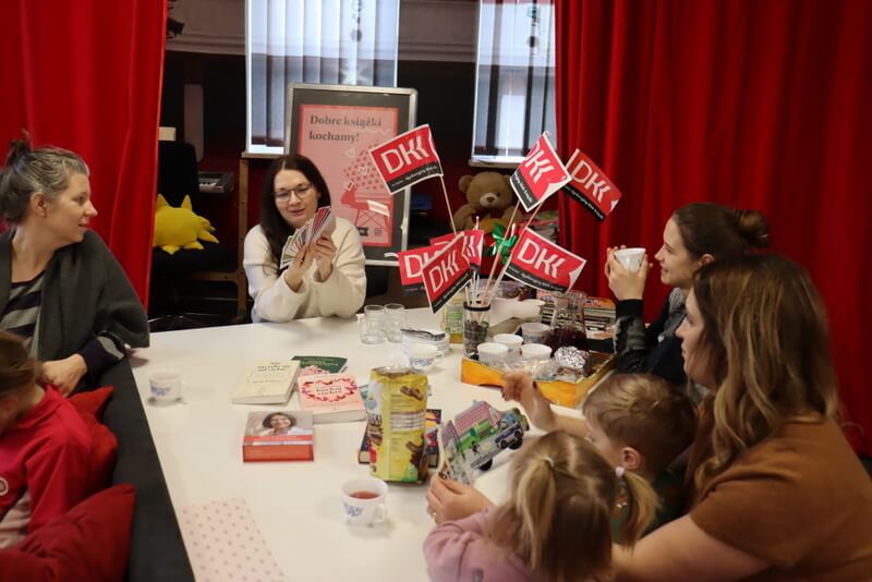 Oddział dla Dzieci. Grupa kobiet siedzi przy białym stole w kształcie fortepianu. Na blacie leżą książki, stoją szklaneczki i filiżanki z herbatą. Stoi wazon z różowymi chorągiewkami z logo DKK. W centrum zdjęcia prowadząca trzyma w dłoniach kolorowe karty, opowiada o wydawnictwie. W tle widoczny plakat Dyskusyjnych Klubów Książki. Po bokach kadru czerwone kotary. Między kobietami siedzą dzieci na kolanach mam.