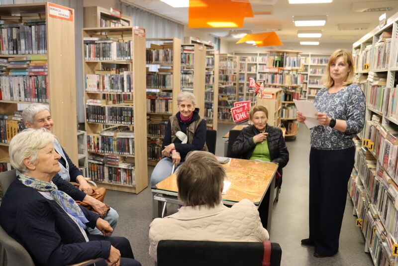 Na pierwszym planie pięć kobiet siedzących na krzesłach przy kwadratowym stoliku, kobieta z prawej strony stoi, w rekach trzyma kartkę,.Na drugim planie stolik , na którym stoi wazon z różowymi chorągiewkami z logo DKK. W głębi regały z książkami. Po lewej i prawej stronie pomieszczenia regały z książkami.