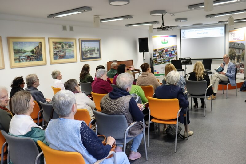 Na pierwszym planie osoby siedzące na krzesłach ustawionych w rzędach, zwrócone przodem do prelegenta widocznego na drugim planie . Prelegent z mikrofonem, siedzi przy okrągłym stoliku, za nim dwa banery, między nimi ekran , z lewej strony pod ścianą stoi pianino, za nim stoi kolumna, na ścianie wiszą obrazy.