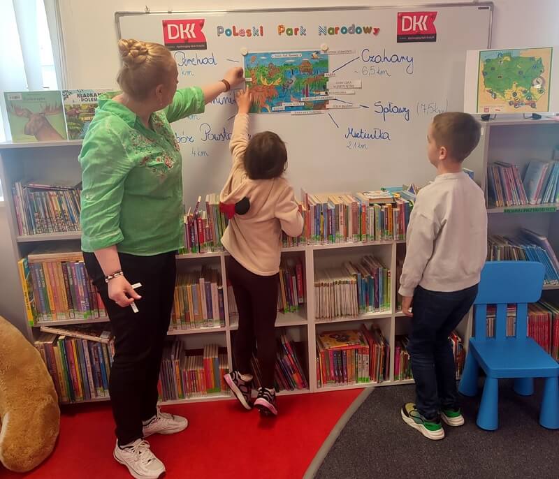 Dzieci wraz z panią stoją przy tablicy zwrócone w kierunku plansz edukacyjnych. Tło stanowią regały z książkami.